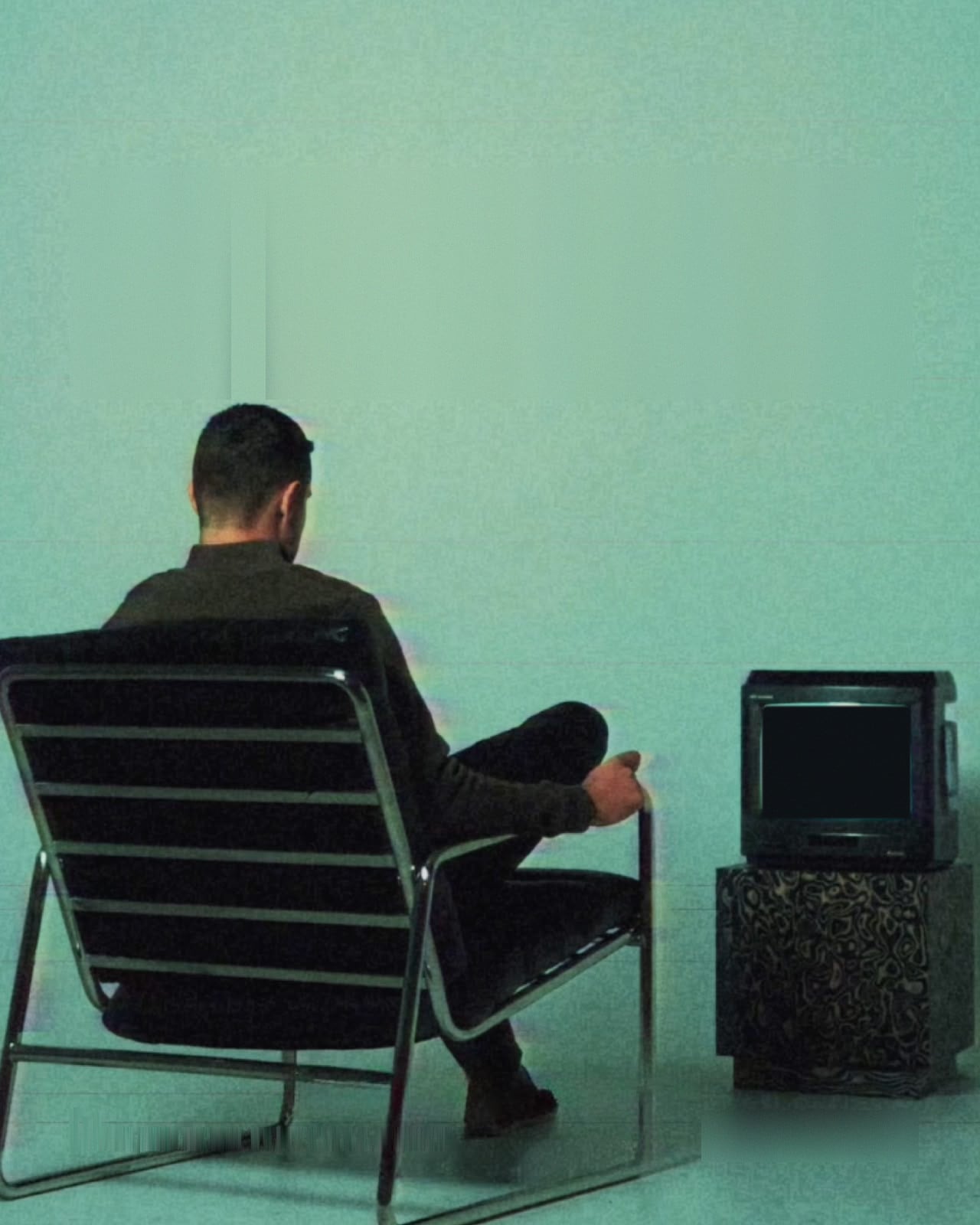 Hamad, seen from behind in a chrome armchair, watching an old CRT television